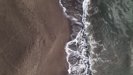 Top-down-view-on-calm-waves-rinsing-on-a-dark-sand-beach