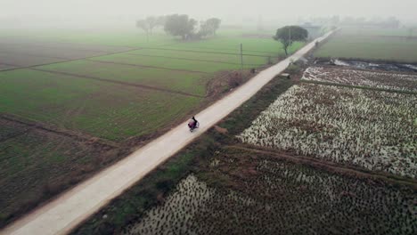 Aerial-shot-of-a-lone-motorcycle-on-a-rural-road-in-Alipur,-Pakistan,-amidst-foggy-fields-at-dawn