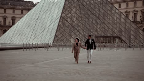 Elegant-couple-running-over-the-empty-place-at-the-Museum-du-Louvre-Pyramid-glas-surface-surrounded-by-the-baroque-royal-residence-buildings-in-Paris-France---modern-lifestyle-and-suits-dating-lovers