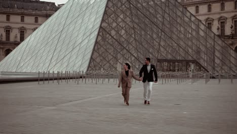 Elegantes-Paar-Läuft-über-Den-Leeren-Platz-Vor-Der-Glaspyramide-Des-Louvre-Museums,-Umgeben-Von-Den-Barocken-Gebäuden-Der-Königlichen-Residenz-In-Paris,-Frankreich---Moderner-Lebensstil-Und-Verabredungen-Für-Liebhaber