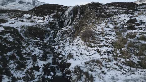 Snow-dusted-waterfall-cascading-down-rocky-terrain-with-mountains-in-background,-shot-in-Skye-Bride-Vail-Fall,-Scotland