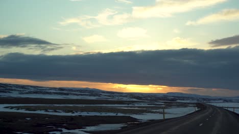 Dusk-settles-over-a-serene-Icelandic-landscape-viewed-from-a-driving-car,-snow-patches-dotting-the-vast-terrain-under-a-dramatic-sky