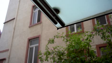 Raindrops-falling-from-a-building's-eave-against-a-blurred-residential-backdrop,-focus-on-water-droplet