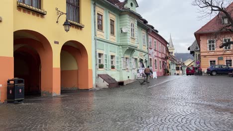 Anciano-Llevando-Un-Gran-Tronco-De-Madera-Durante-Un-Día-Lluvioso-A-Través-De-La-Histórica-Ciudad-De-Sighisoara,-Rumania