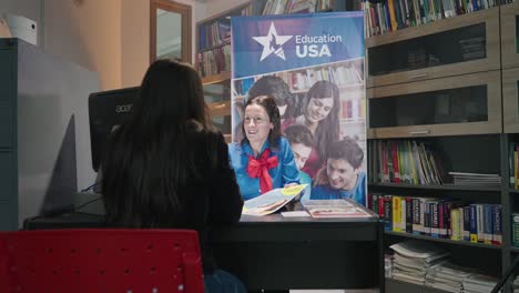 Bewegte-Aufnahme-Einer-Kaukasischen-Frau-In-Uniform,-Die-An-Einem-Schreibtisch-Sitzt-Und-Einer-Anderen-Frau-In-Einem-Büro-Mit-Büchern-Und-Einem-Schild-Mit-Der-Aufschrift-„Education-USA“-Informationen-Gibt