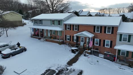 Snow-covers-a-residential-area-with-two-story-homes,-bare-trees,-two-parked-cars,-and-a-clear-driveway