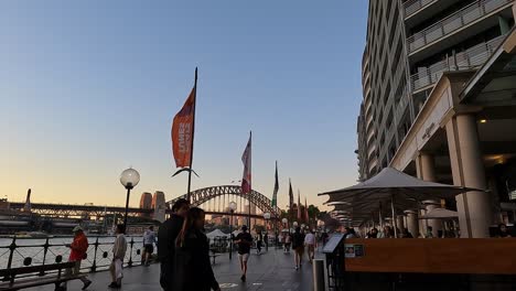 Hora-Dorada-En-El-Vibrante-Puerto-De-Sydney-Con-Peatones-Y-El-Icónico-Puente-Del-Puerto-A-La-Vista,-Tonos-Cálidos