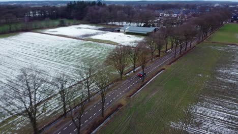 Aerial-above-scenic-road-through-the-snow-covered-agricultural-fields-and-farms-with-car-driving-to-Bergeijk-town