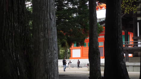 Visitors-walk-by-the-Red-Konpon-Daito-pagoda-in-Koyasan,-surrounded-by-towering-trees,-zoom-out