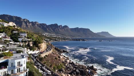 Cape-Town,-South-Africa-aerial-over-a-coastline-road,-with-Camps-Bay-in-the-distance