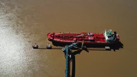 A-red-tanker-ship-docking-on-a-river-during-daytime,-sunny,-aerial-view