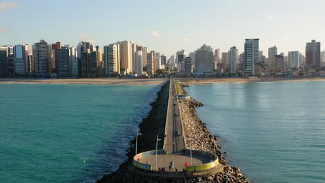Aerial-view-of-footbridge-with-people-walking-around,-from-the-sea-to-the-city-with-many-buildings-in-the-background,-Fortaleza,-Ceara,-Brazil