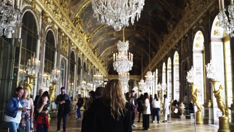 Blonde-Frau-In-Einer-Schwarzen-Jacke-Zeichnet-Ihre-Sicht-Auf,-Während-Sie-Mit-Ihrem-Telefon-Durch-Den-Spiegelsaal-Im-Schloss-Versailles-In-Paris-Frankreich-Geht,-Mit-Kronleuchtern-Und-Wänden-Voller-Historischer-Gemälde-Und-Goldener-Ornamente