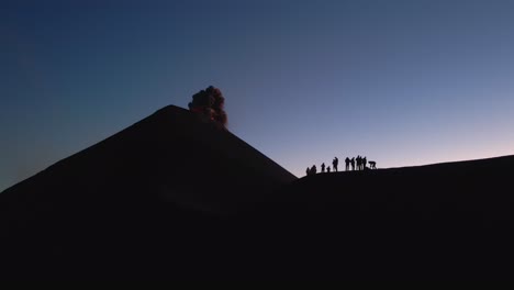 Eine-Filmische-Luftaufnahme-Einer-Gruppe-Auf-Dem-Grat-Des-Vulkans-Fuego-Zeigt,-Wie-Dieser-Ausbricht-Und-Rote-Lava-Und-Asche-Ausspuckt.