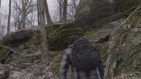 A-hiker-with-a-black-backpack-and-a-full-beard-climbs-up-ti-rocky-hillside-among-the-mossy-cliffs-and-barren-trees-in-ohio-on-a-gray-winter-day