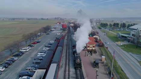 An-Aerial-View-of-a-Train-Station,-with-a-Steam-Passenger-Train,-Pulling-into-the-Station,-Blowing-Smoke,-on-a-Partially-Sunny-Day