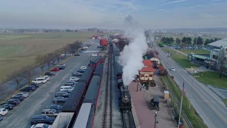 Una-Vista-Aérea-De-Una-Estación-De-Tren,-Con-Un-Tren-De-Pasajeros-A-Vapor,-Entrando-En-La-Estación,-Expulsando-Humo,-En-Un-Día-Parcialmente-Soleado.