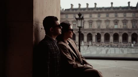 Couple-with-black-sunglases-leaning-on-the-wall-enjoying-the-view-on-the-empty-place-around-the-glas-pyramides-of-Museum-du-Louvre-with-it-borque-buildings-and-fountain-around-in-Paris-France