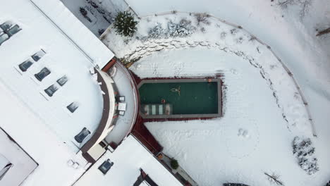 Toma-Aérea-De-Un-Lujoso-Hotel-Y-Spa-En-Medio-De-Las-Montañas,-Cubierto-De-Nieve-Invernal-Con-Una-Increíble-Piscina-Con-Agua-Humeante,-Gente-Relajándose-Después-De-Esquiar