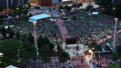 Aerial-view-of-people-enjoying-evening-in-park,-sitting-on-lawn-and-watching-large-screen