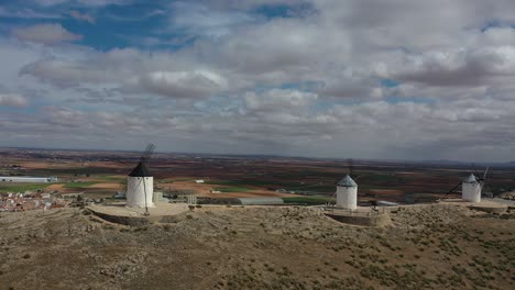 Seitlicher-Flug-Mit-Einer-Drohne-Auf-Einem-Hügel,-Der-Drei-Alte-Windmühlen-Visualisiert,-Die-Von-Menschen-Besucht-Werden,-Mit-Einem-Hintergrund-Von-Ackerland-Und-Einer-Gewissen-Bevölkerung-An-Einem-Frühlingstag-Mit-Einem-Himmel-Mit-Wolken,-Toledo,-Spanien