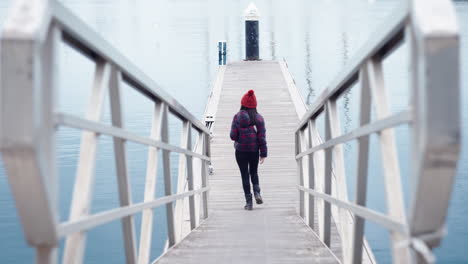 Una-Mujer-Camina-Hacia-El-Puerto-Con-Un-Gorro-Rojo-Y-Una-Chaqueta-A-Cuadros,-Con-Aguas-Cristalinas-Como-Telón-De-Fondo-Mientras-Los-Pájaros-Vuelan-Con-Gracia-En-Primer-Plano,-Lo-Que-Se-Suma-A-La-Pintoresca-Escena.