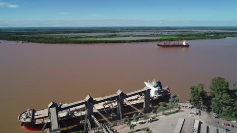 Cargo-ships-and-grain-terminal-on-a-muddy-river,-clear-sky,-aerial-view