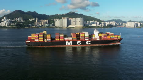 wide-angle-shot-of-a-giant-container-vessel-passing-in-front-of-tropical-island-with-urban-sprawl