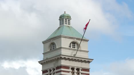 The-top-of-the-building-exterior-of-Central-Fire-Station-with-national-flag-waving-against-blue-sky-and-fast-moving-clouds,-museum-of-the-Singapore-Civil-Defence-Force-on-hill-street,-close-up-shot