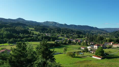 drone-flight-in-ascent-looking-at-the-treetops-in-the-Basque-Cantabra-area-over-a-rural-town-with-farmland-and-lush-oak-forests-and-mountains-on-an-afternoon-with-blue-sky-in-summer-and-green-grass