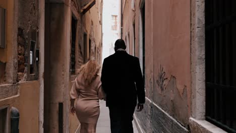 Una-Elegante-Pareja-De-Raza-Mixta-Caminando-Por-Las-Estrechas-Calles-De-Venecia,-Italia,-Entre-Las-Típicas-Casas-Antiguas-Para-Explorar-La-Ciudad-Mientras-Viaja,-Vista-Desde-Atrás