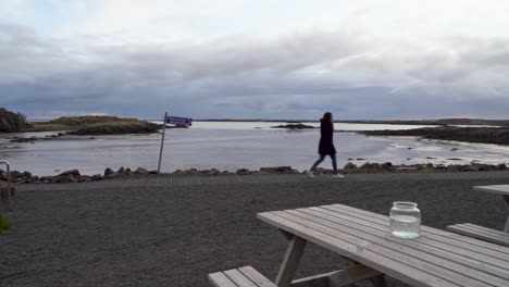 Woman-walking-by-a-serene-Icelandic-coast,-cloudy-skies,-reflective-water,-with-picnic-tables-in-foreground