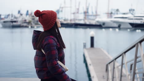 Asian-woman-rests-by-the-water-at-Valencia-port-Spain,-contemplating-as-birds-perch-nearby,-their-presence-adding-to-the-serene-scene-with-a-red-beanie,-harmonizing-with-her-matching-chequered-jacket