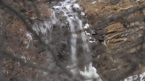 Through-wooden-branches,-focus-rack-view,-cascading-water-over-rocky-cliff