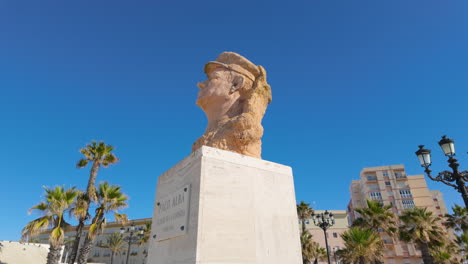 Bust-of-Paco-Alba,-known-as-the-"Father-of-Chirigota,"-displayed-on-a-white-pedestal,-with-palm-trees-and-a-clear-blue-sky-in-the-background,-suggesting-a-warm,-sunny-location,-likely-in-Cádiz