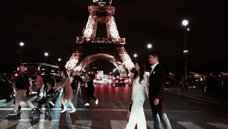 Elegante-Pareja-De-Cabello-Oscuro-Con-Traje-Blanco-Y-Negro-Caminando-Por-La-Calle-En-Pont-D&#39;lena-Frente-A-La-Famosa-Torre-Eiffel-Por-La-Noche-Mientras-Toda-La-Gente-Y-Los-Semáforos-Toman-Algunas-Fotos