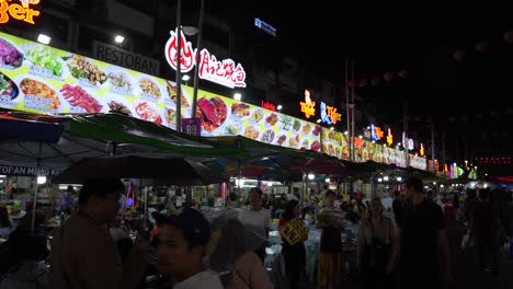 Street-Food-In-Kuala-Lumpur:-Lebhafte-Atmosphäre-Eines-Nachtmarkts-Mit-Hellen-Neonschildern,-Die-Für-Verschiedene-Lebensmittel-Werben,-über-Einer-Belebten-Straße-Mit-Essensständen-Und-Einer-Geschäftigen-Menschenmenge