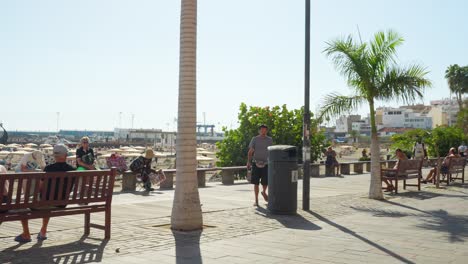 Tourists-and-locals-walking-and-sitting-by-the-bay-walk-in-Playa-De-Las-Americas-in-Tenerife,-dynamic-panning-shot