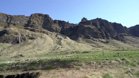 Lush-green-Icelandic-landscape-with-rugged-cliffs-and-a-small-waterfall-under-clear-blue-skies