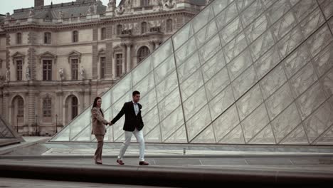 Elegantes-Paar-Spaziert-Auf-Der-Niedrigen-Mauer-Der-Glaspyramide-Des-Museum-Du-Louvre,-Umgeben-Von-Den-Barocken-Gebäuden-Der-Königlichen-Residenz-In-Paris,-Frankreich---Moderner-Lebensstil-Und-Verabredungen-Für-Liebhaber