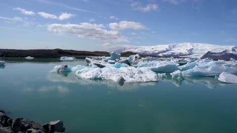 Vibrant-blue-icebergs-floating-in-Jokulsarlon-Glacier-Lagoon-with-Icelandic-mountains-in-the-background,-clear-sky