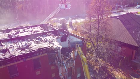 Large-excavator-demolishing-building-that-was-destroyed-by-fire-and-firetruck-aerial-platform-in-Blainville,-Canada