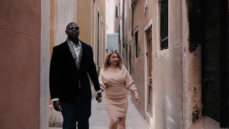 Una-Elegante-Pareja-De-Raza-Mixta-Caminando-Por-Las-Estrechas-Calles-De-Venecia,-Italia,-Entre-Las-Típicas-Casas-Antiguas-Para-Explorar-La-Ciudad-Mientras-Viaja.