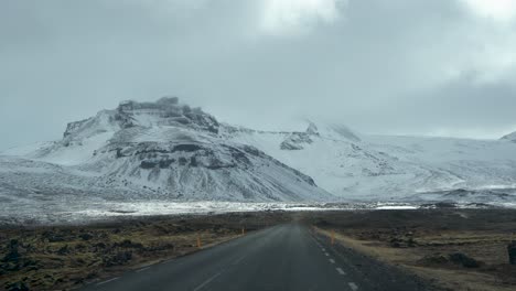 Blick-Des-Fahrers-Auf-Eine-Offene-Straße,-Die-Zu-Einem-Schneebedeckten-Berg-In-Island-Führt,-Darüber-Bewölkter-Himmel