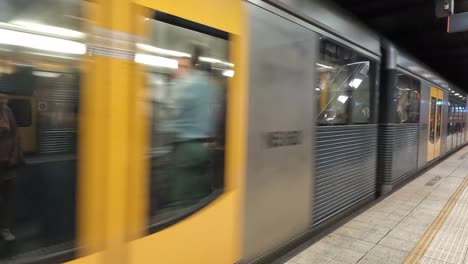 Blurry-motion-of-yellow-train-passing-by-a-platform-with-people-waiting,-Sydney-station
