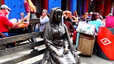 Escultura-De-Niña-De-Galway-En-Quay-Street-Mientras-Los-Turistas-Disfrutan-En-El-Bar-De-Quays