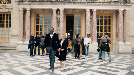 Elegant-Gekleidetes-Paar-Gemischter-Abstammung,-Das-An-Einem-Windigen-Tag-Aus-Dem-Hauptgebäude-Des-Berühmten-Schlosses-Versailles-In-Paris,-Frankreich,-Auf-Den-Cour-Royale-Geht-Und-Die-Majestätische-Barocke-Architektur-Offenbart