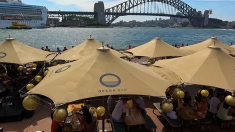 Bar-De-La-Ópera-De-Sydney-Iluminado-Por-El-Sol-Con-Clientes,-Cruceros-Y-El-Puente-Del-Puerto-Al-Fondo,-Durante-El-Día