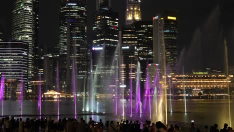 Crowds-gathered-at-the-Marina-bay-sands-event-plaza,-watching-SPECTRA-light-and-water-show-in-the-evening-with-downtown-cityscape-in-the-background,-timelapse-static-shot