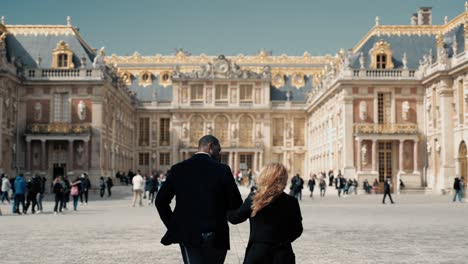 Una-Pareja-De-Raza-Mixta-Vestida-De-Negro-Caminando-Por-El-Cour-Royale-Hasta-El-Edificio-Principal-Del-Famoso-Castillo-Barroco-De-Versalles-En-París,-Mientras-Hay-Turistas-En-Un-Día-Soleado,-Tomado-Desde-Atrás